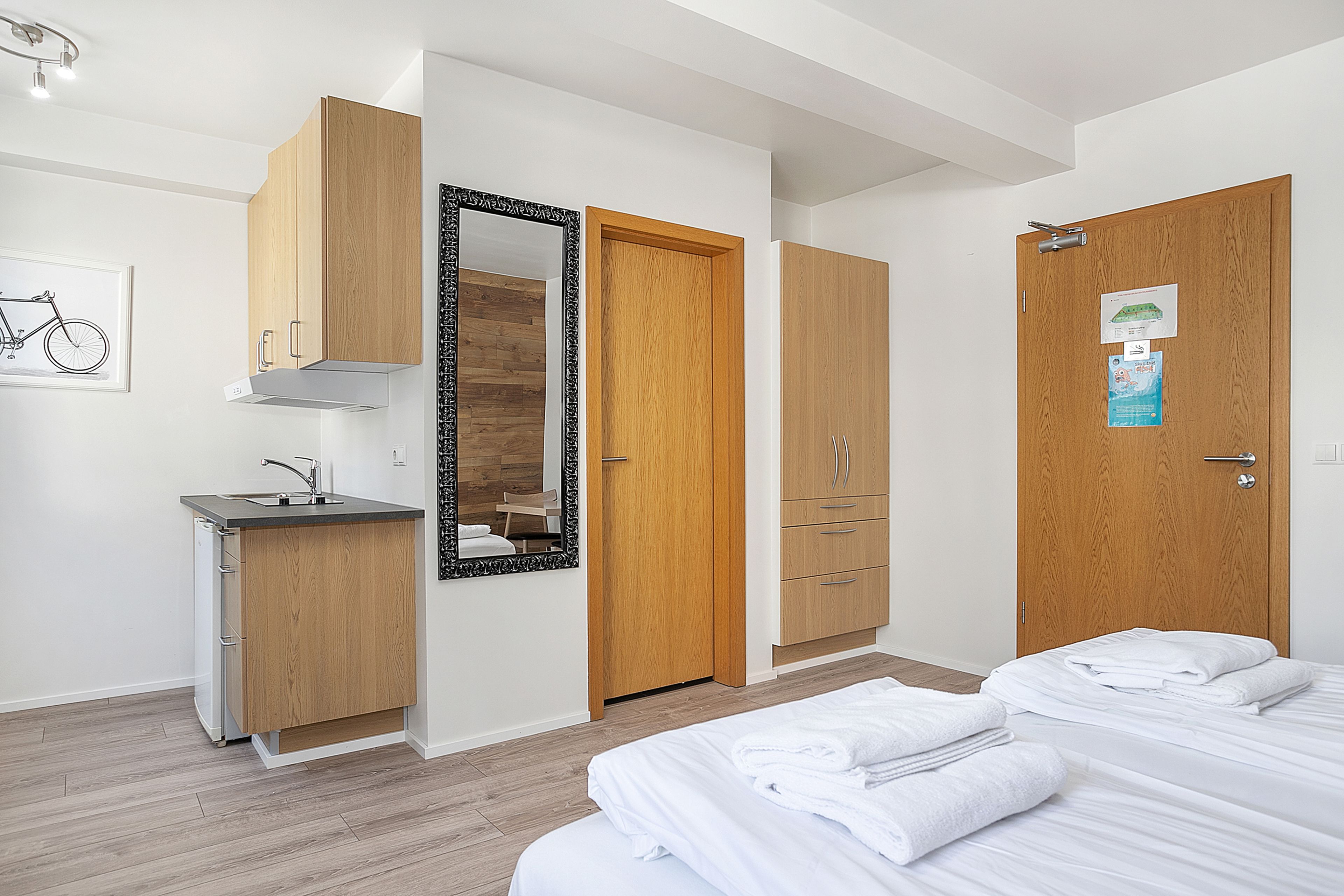A modern hotel room with a wooden wardrobe, a small kitchenette, a large mirror, and a bed with neatly folded white towels.