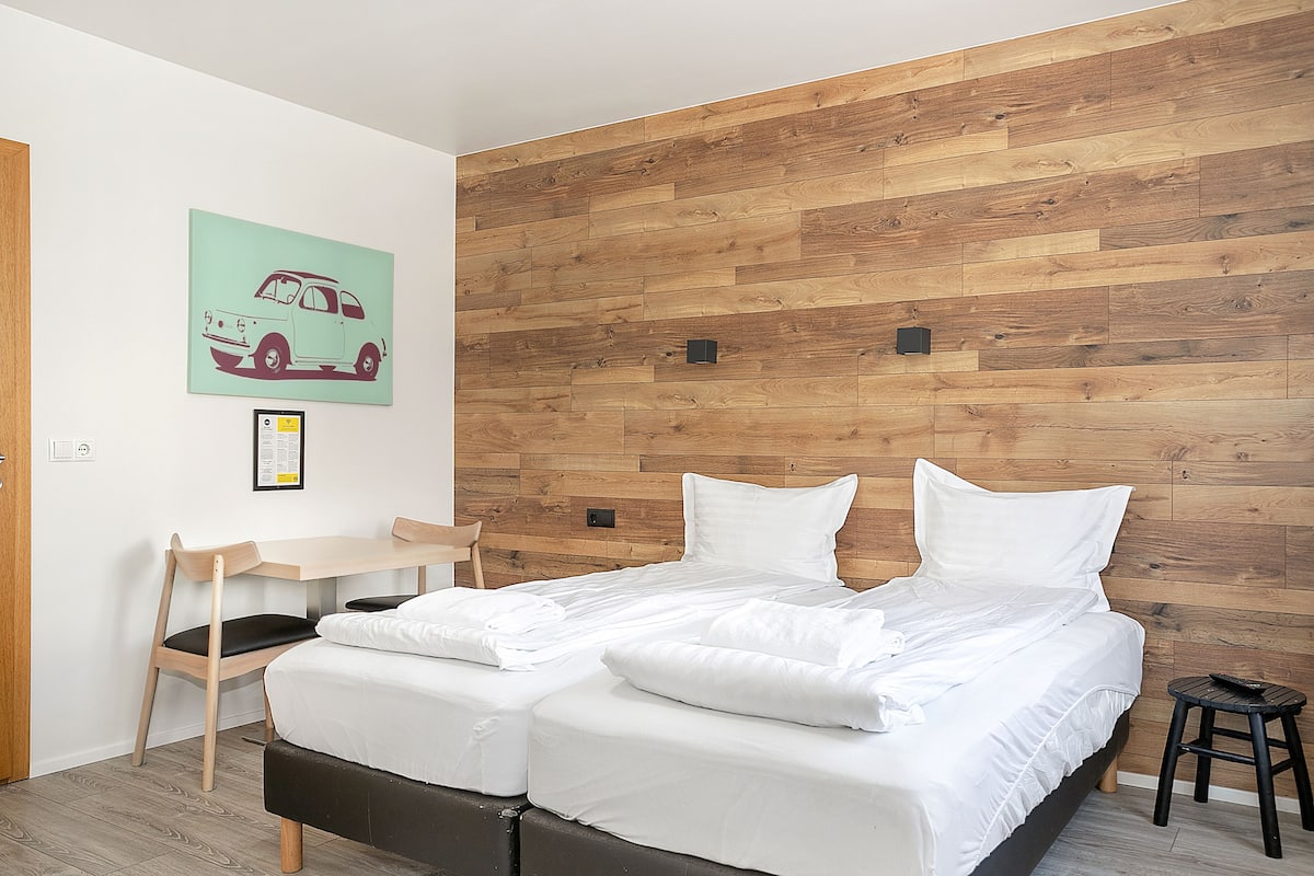 Quarto moderno com cabeceira de madeira, cama com roupa de cama branca, escrivaninha pequena com cadeira e quadro na parede de um carro.