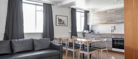 Un salon moderne comprenant un canapé, une table à manger et des chaises, un coin cuisine avec des meubles en bois et de grandes fenêtres dotées de rideaux.