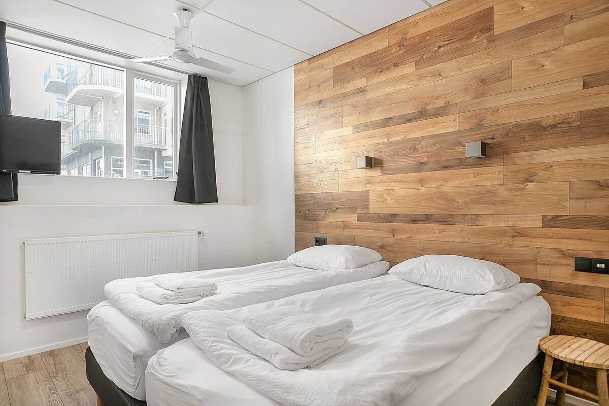 Quarto de hotel moderno com uma cama grande, uma parede de destaque em madeira, uma janela vista para edifícios e um ventilador de teto.