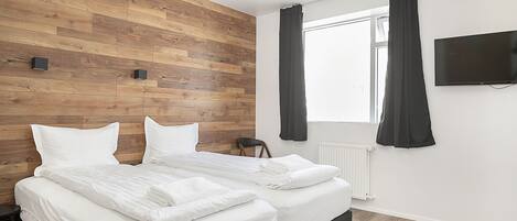 A bedroom with two beds, a wooden wall, a window with curtains, a radiator, and a TV on the wall.