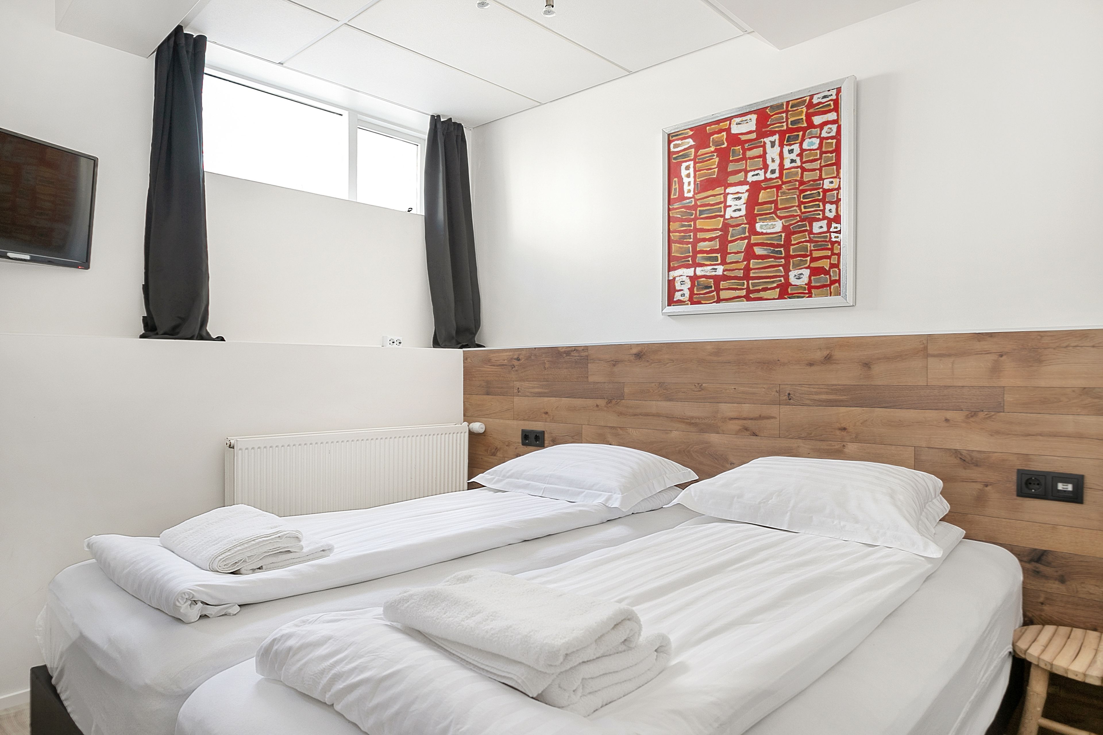 Une chambre d’hôtel avec deux lits, une tête de lit en bois, une fenêtre avec des rideaux noirs et un tableau abstrait rouge accroché au mur.
