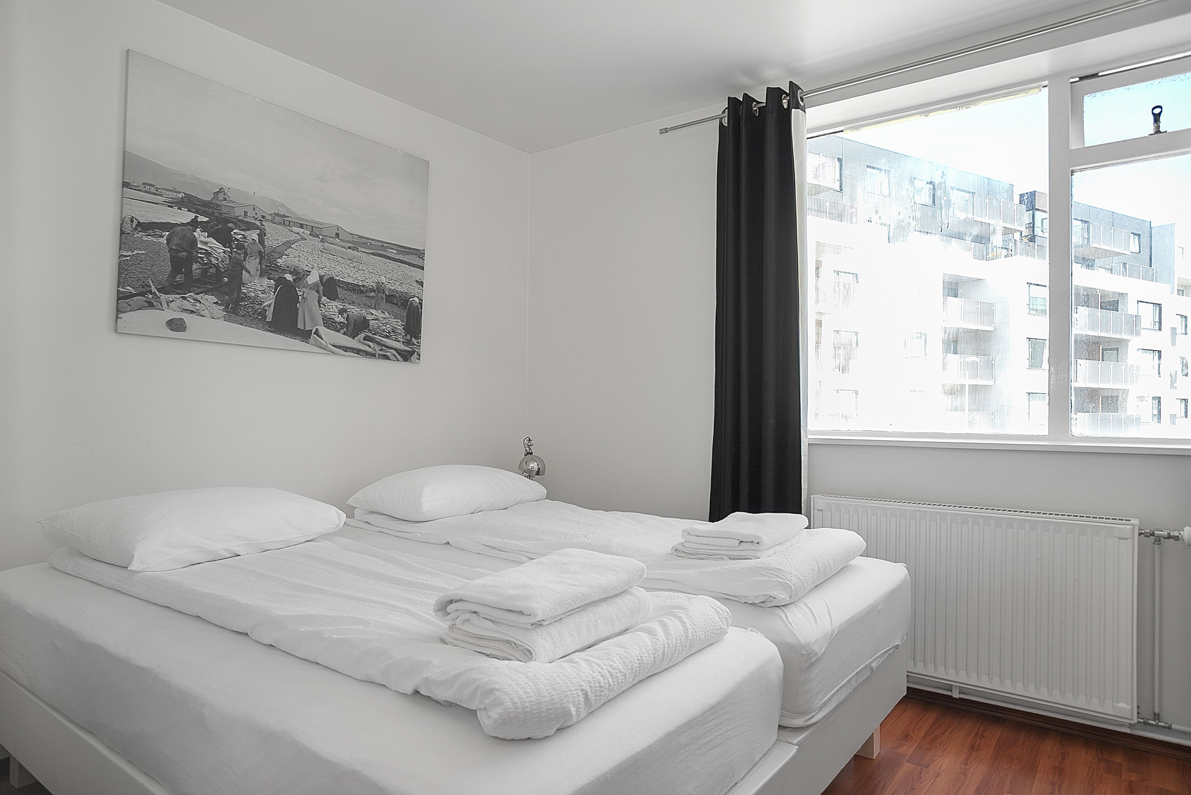 Une chambre d’hôtel avec deux lits, du linge de lit blanc, un rideau noir, un radiateur blanc et une photographie en noir et blanc accrochée au mur.