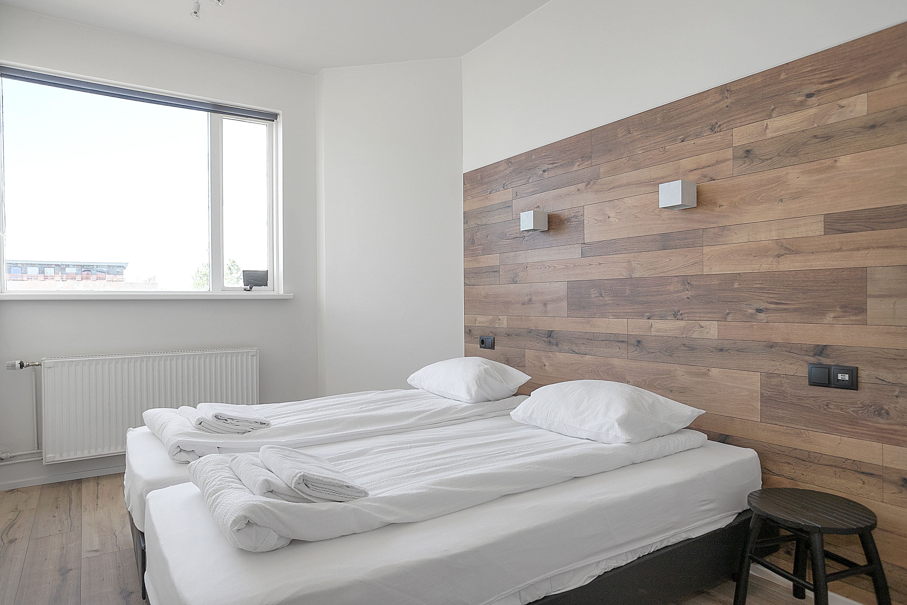 A modern hotel room with two beds, a wooden accent wall, and a large window.