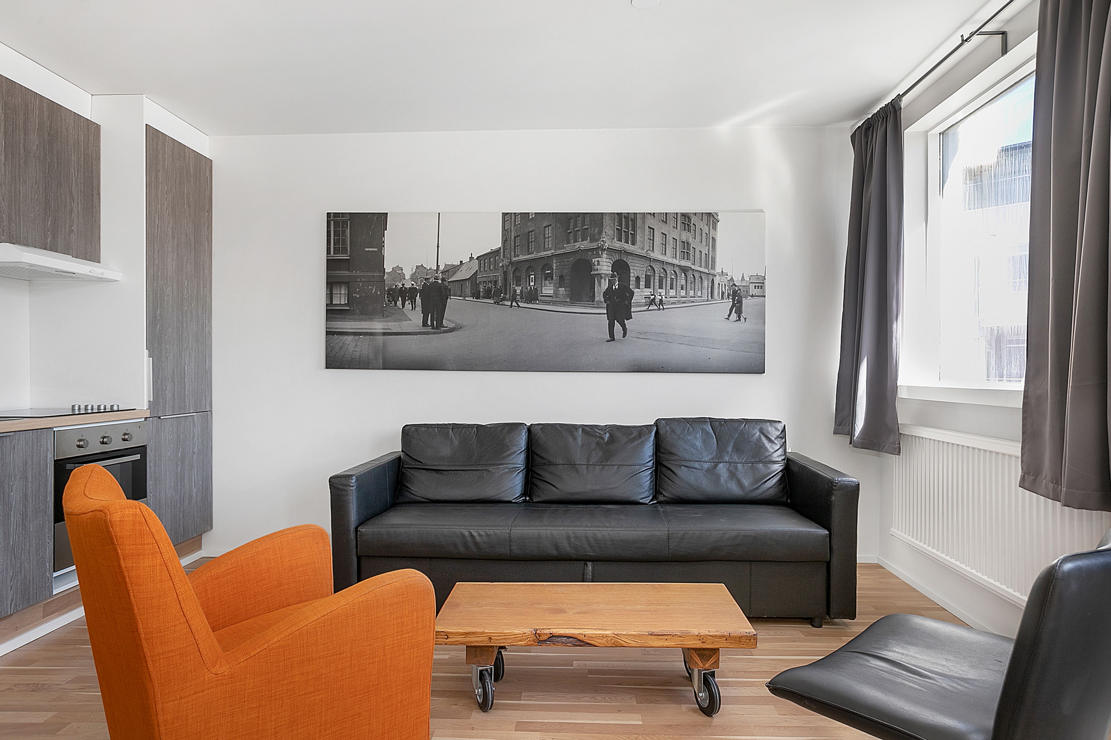 Un salon moderne avec un canapé en cuir noir, un fauteuil orange et une table basse en bois. Une photographie en noir et blanc représentant une scène de rue est accrochée au mur.