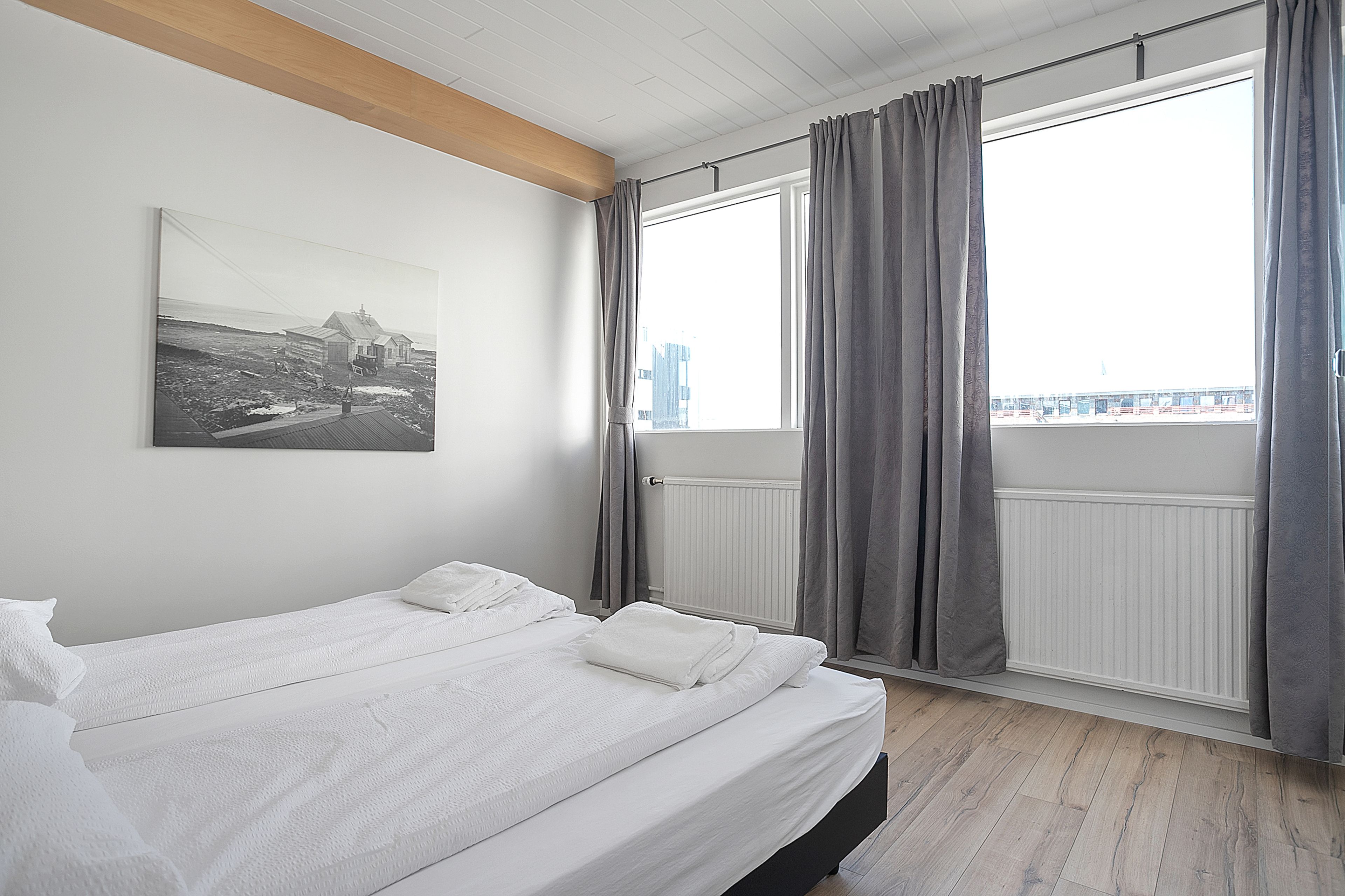 A bedroom with two beds, a large window with curtains, a radiator, and a city view.