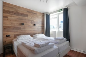 Habitación de hotel con dos camas, paneles de madera en la pared, una ventana con cortinas y vistas a edificios.
