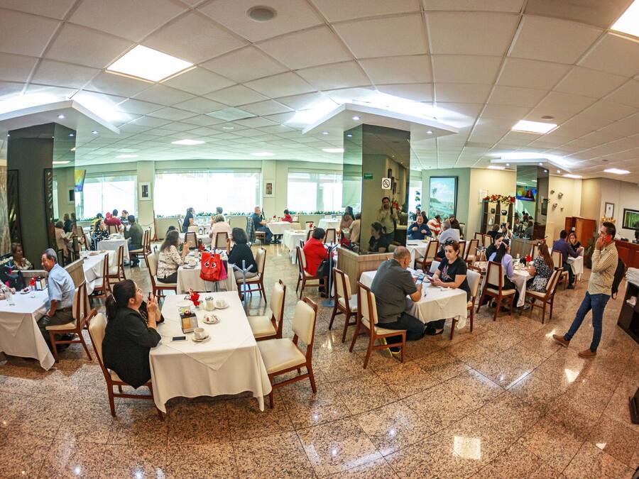 A spacious dining area with multiple tables and chairs, large windows, and a high ceiling with recessed lighting.