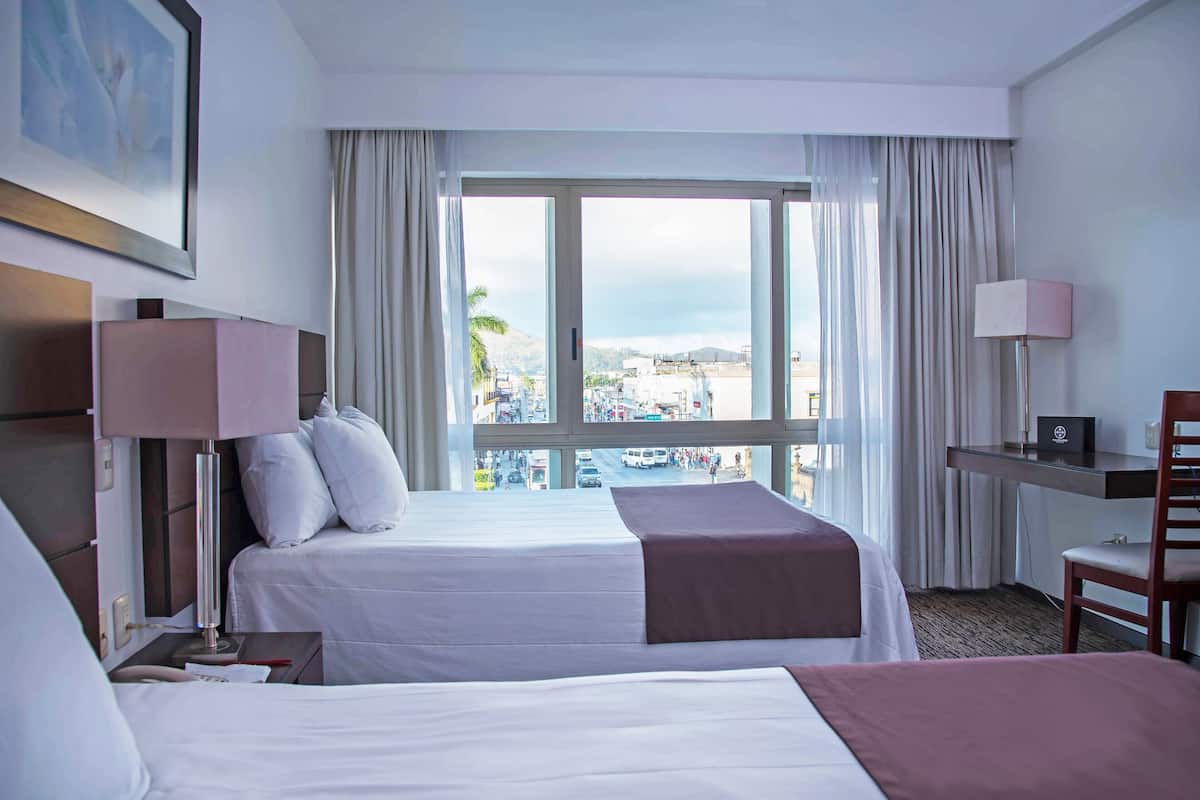 A hotel room with two beds, a desk, a chair, a lamp, a window with curtains, and a view of the city.