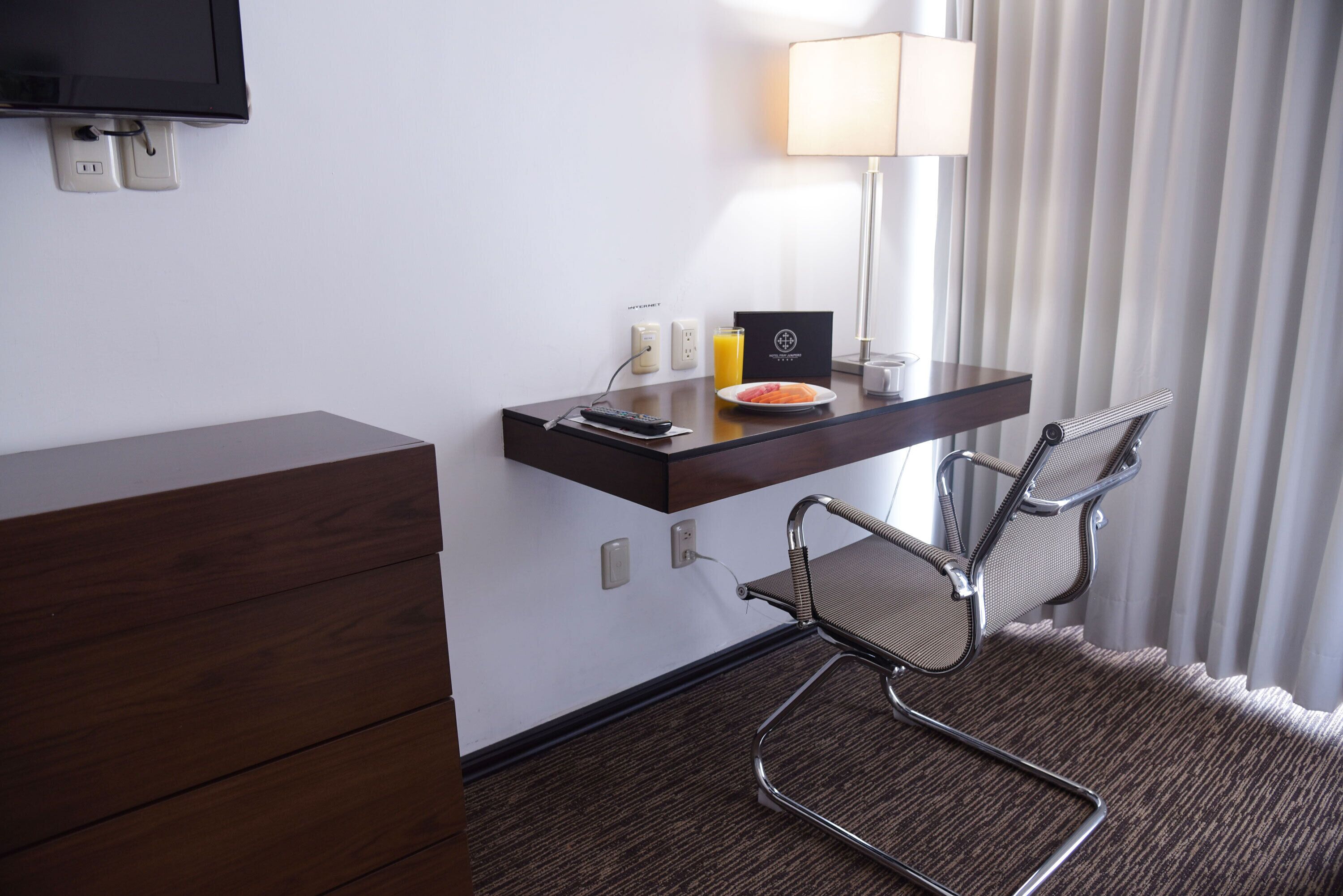 A hotel room with a wooden dresser, a desk with a lamp, a plate of food, and two mesh chairs.
