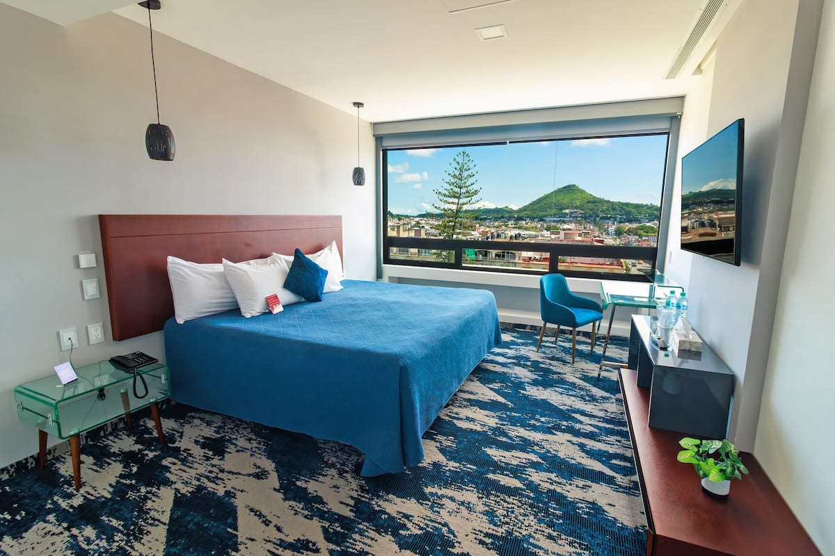 A hotel room with a large bed, a glass side table, a desk with a chair, a TV, and a view of a cityscape through a large window.