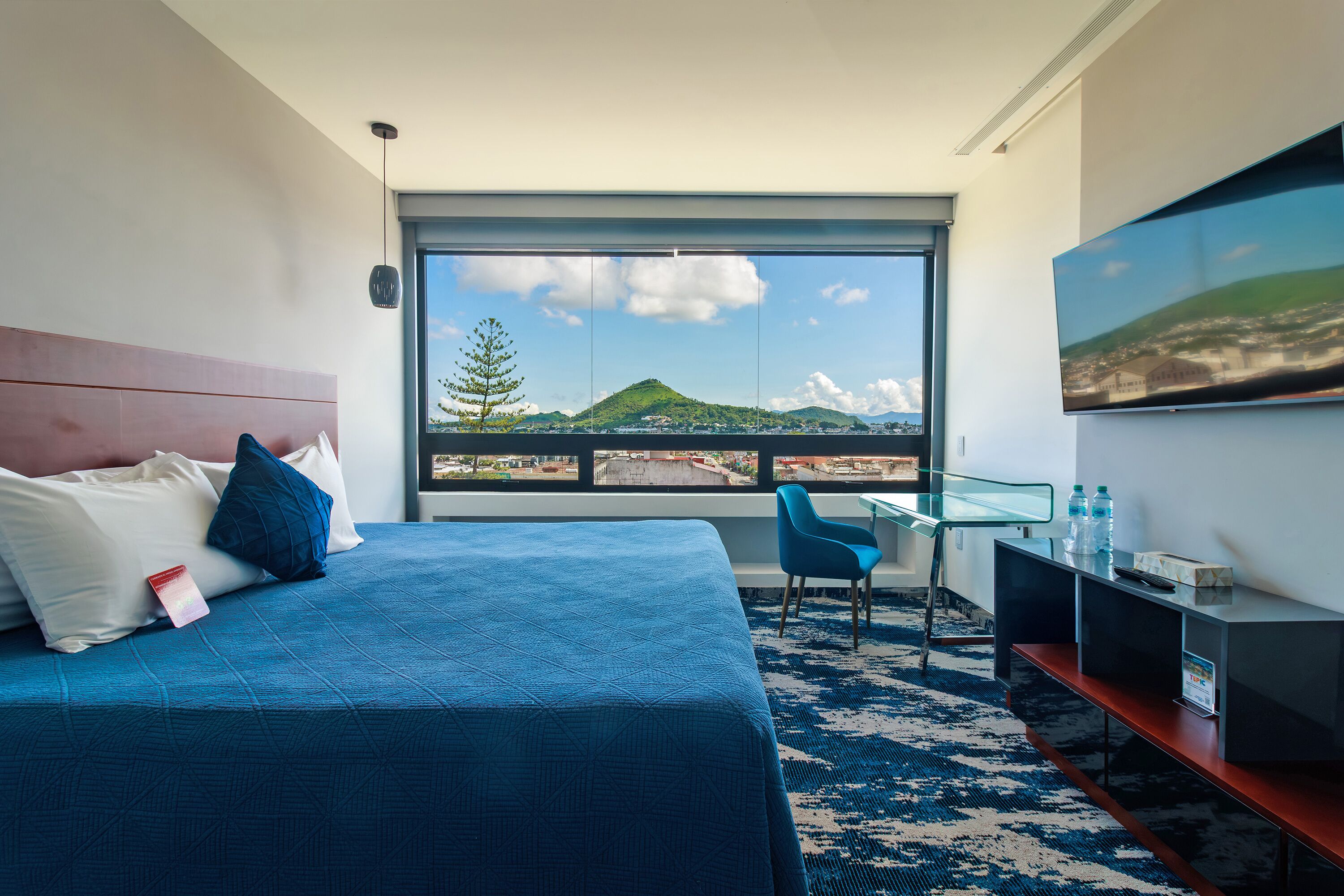 Habitación de hotel moderna con una cama grande, un escritorio y vistas a la montaña y al paisaje urbano a través de un ventanal amplio.