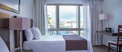 Habitación de hotel con dos camas, un escritorio, una silla, una lámpara y un ventanal con vistas a la ciudad.