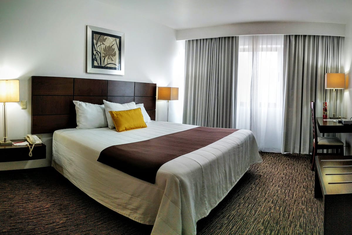A hotel room with a large bed, a headboard, two bedside lamps, a flat-screen TV, a desk with a chair, and a window with curtains.
