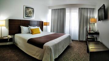 Une chambre d’hôtel avec un grand lit, une tête de lit, deux lampes de chevet, un téléviseur à écran plat, un bureau avec une chaise et une fenêtre avec des rideaux.
