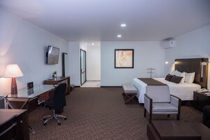 A hotel room with a desk, chair, bed, bedside table, lamp, and artwork.