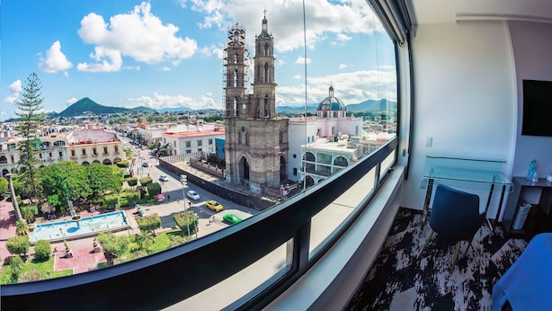 Vista desde una ventana que da a un paisaje urbano con una iglesia destacada, una piscina y edificios circundantes.