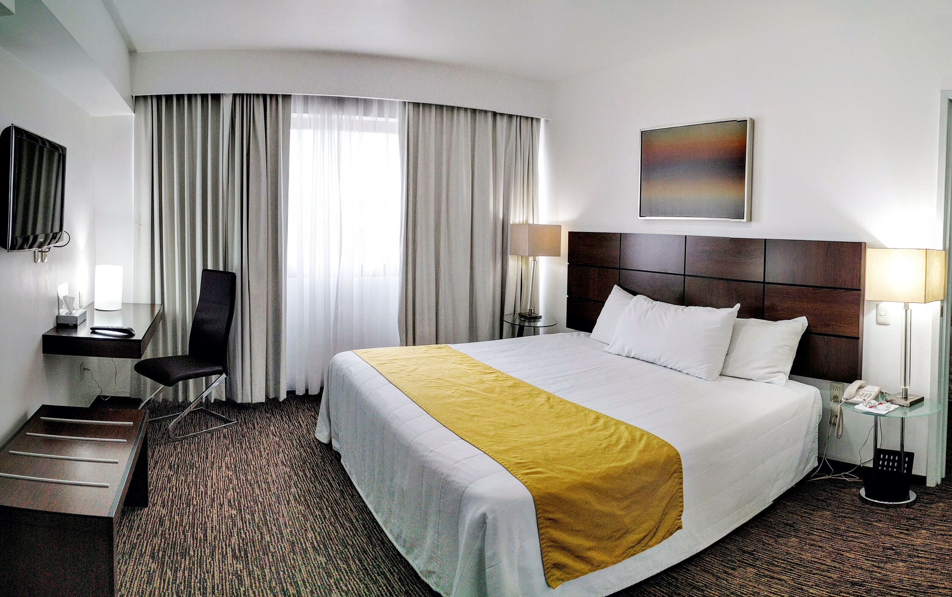 A hotel room with a large bed, a desk with a chair, a TV, and a painting on the wall.