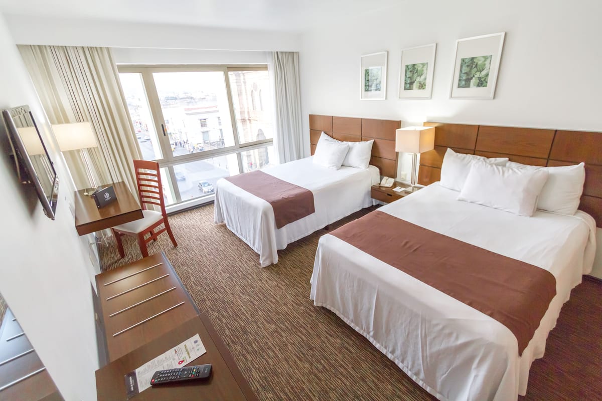A hotel room with two beds, a desk, a chair, a TV, and a view of the city.