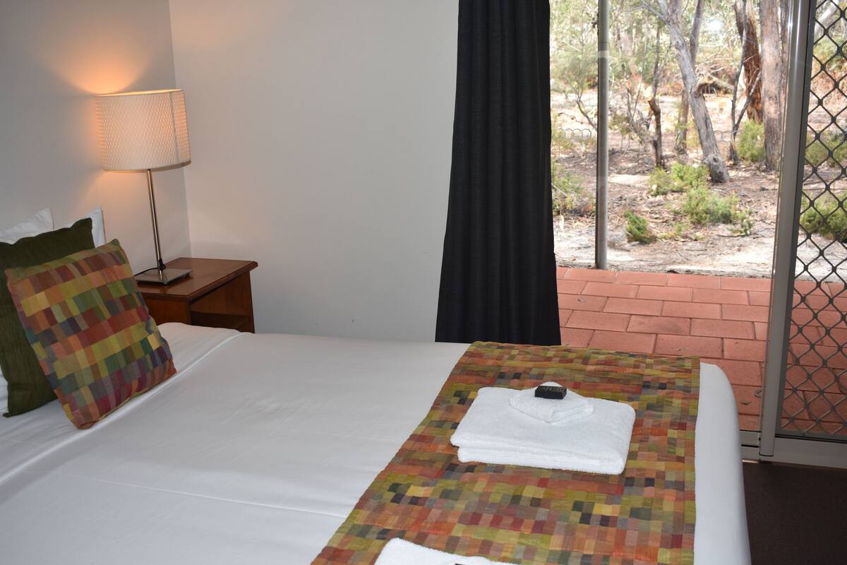 Ein Hotelzimmer mit Bett, Nachttisch, Lampe und Blick ins Freie.