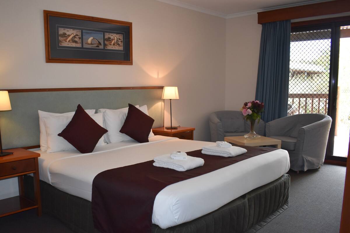 Ein Hotelzimmer mit einem großen Bett, zwei Nachttischen inklusive Lampen, einem Sessel, einem kleinen Tischchen mit einer Vase und einem Balkon mit Blick.