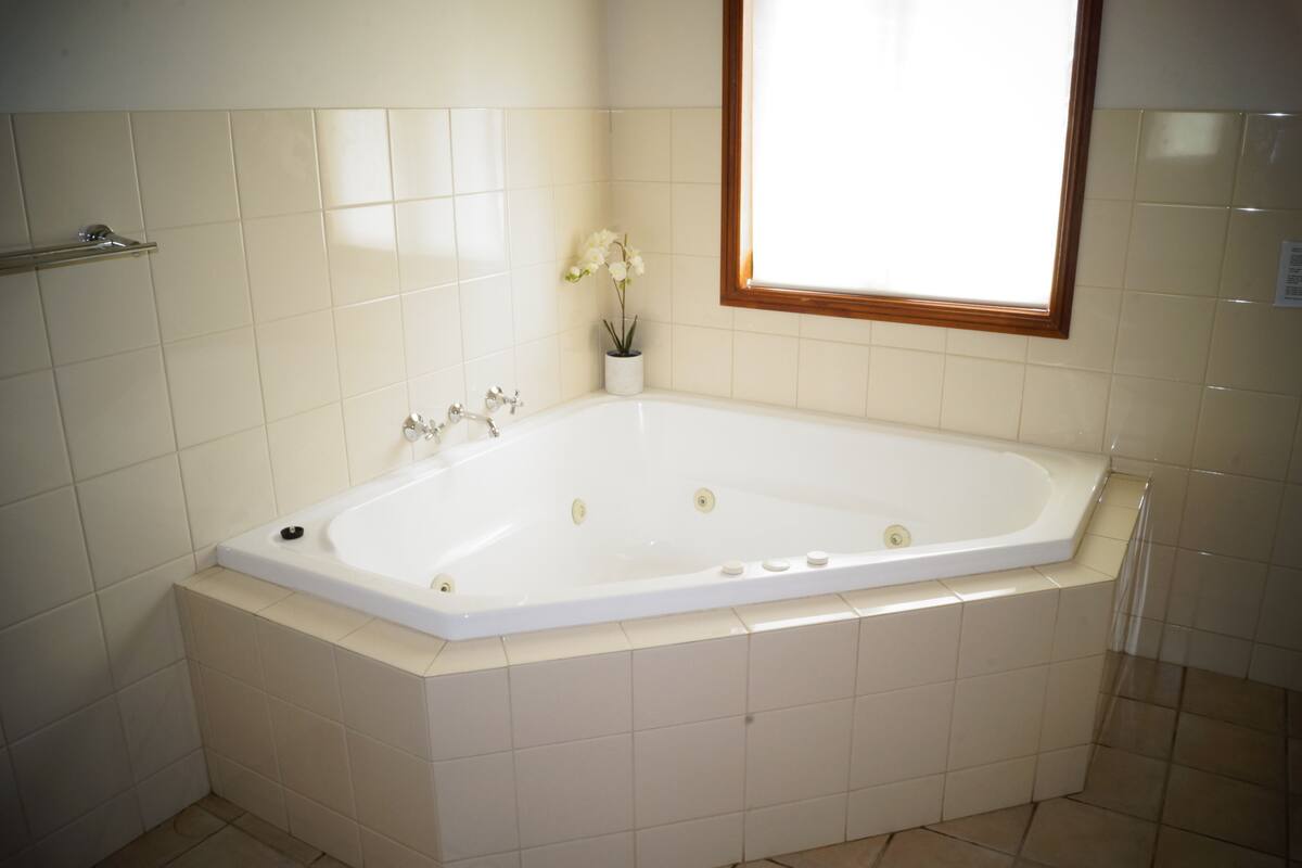 Eine große, rechteckige Badewanne mit Holzrahmen vor dem Fenster, weißen Fliesenwänden und einer kleinen Topfpflanze an der Seite.