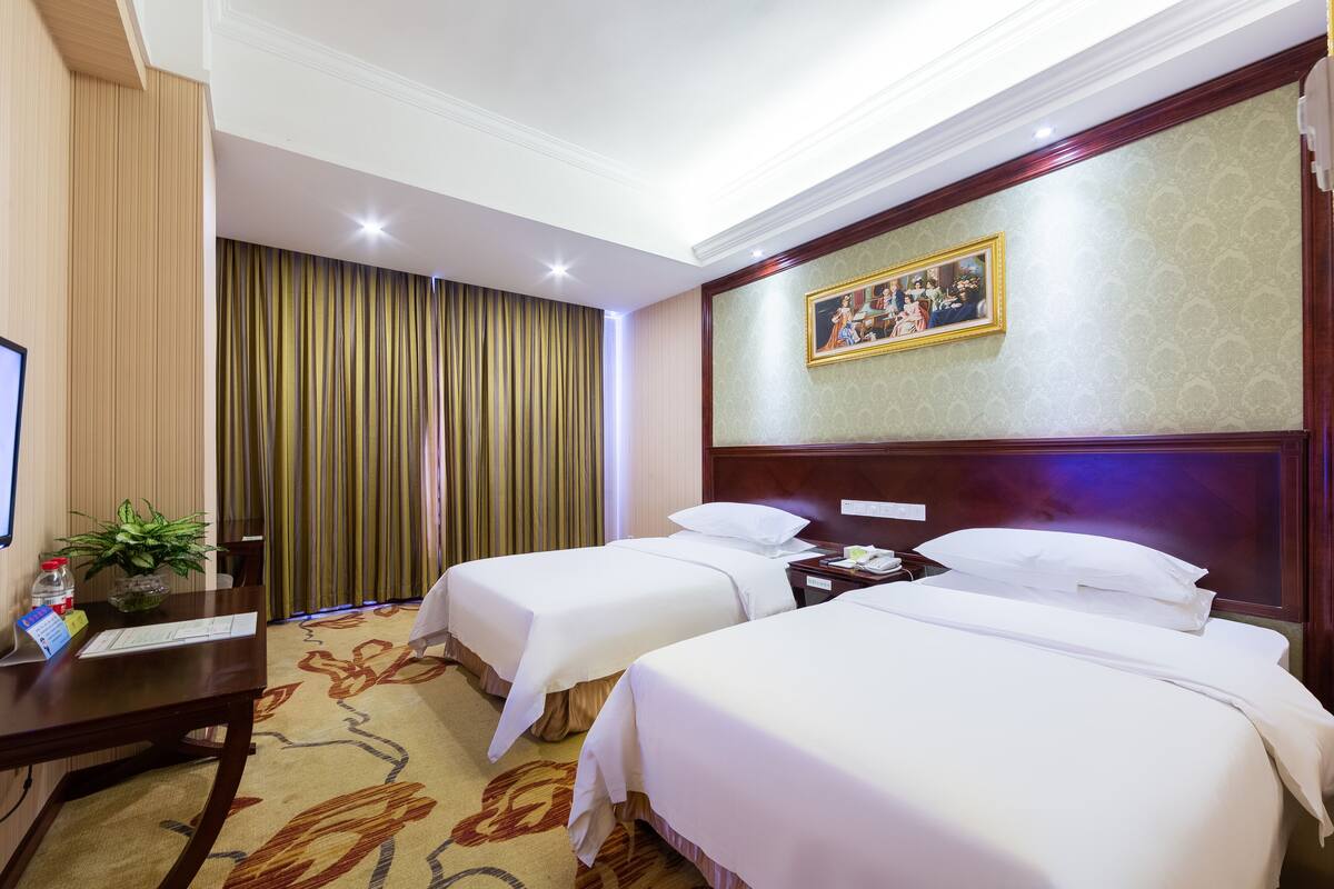 A hotel room with two beds, a desk with a plant, a painting on the wall, and a carpeted floor.