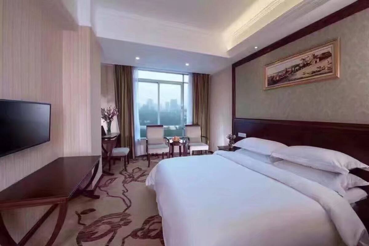 A hotel room with a large bed, a flat-screen TV, a desk, and a view of the city through the window.