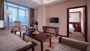 Habitación de hotel con una cama grande, un televisor montado en la pared, una mesa de comedor con sillas y un amplio ventanal con cortinas.
