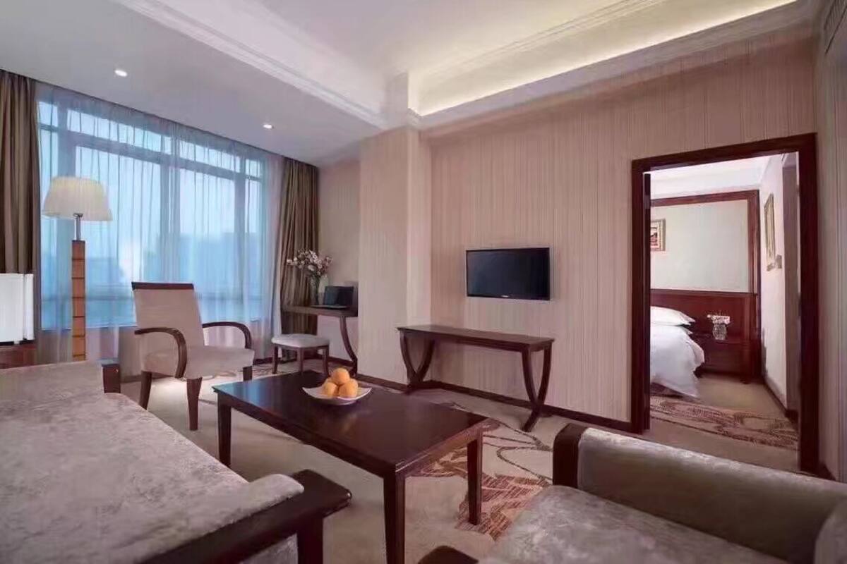 A hotel room with a large bed, a TV mounted on the wall, a dining table with chairs, and a large window with curtains.