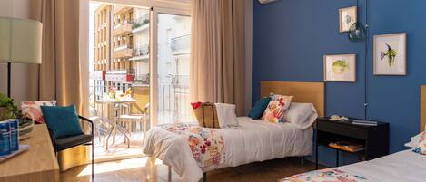 A hotel room with two beds, a desk, a chair, a small table, and a view of the outside balcony.