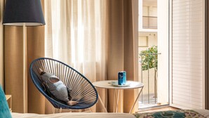 A hotel room with a bed, a blue chair, a small table with a can, and a view of the outside balcony.