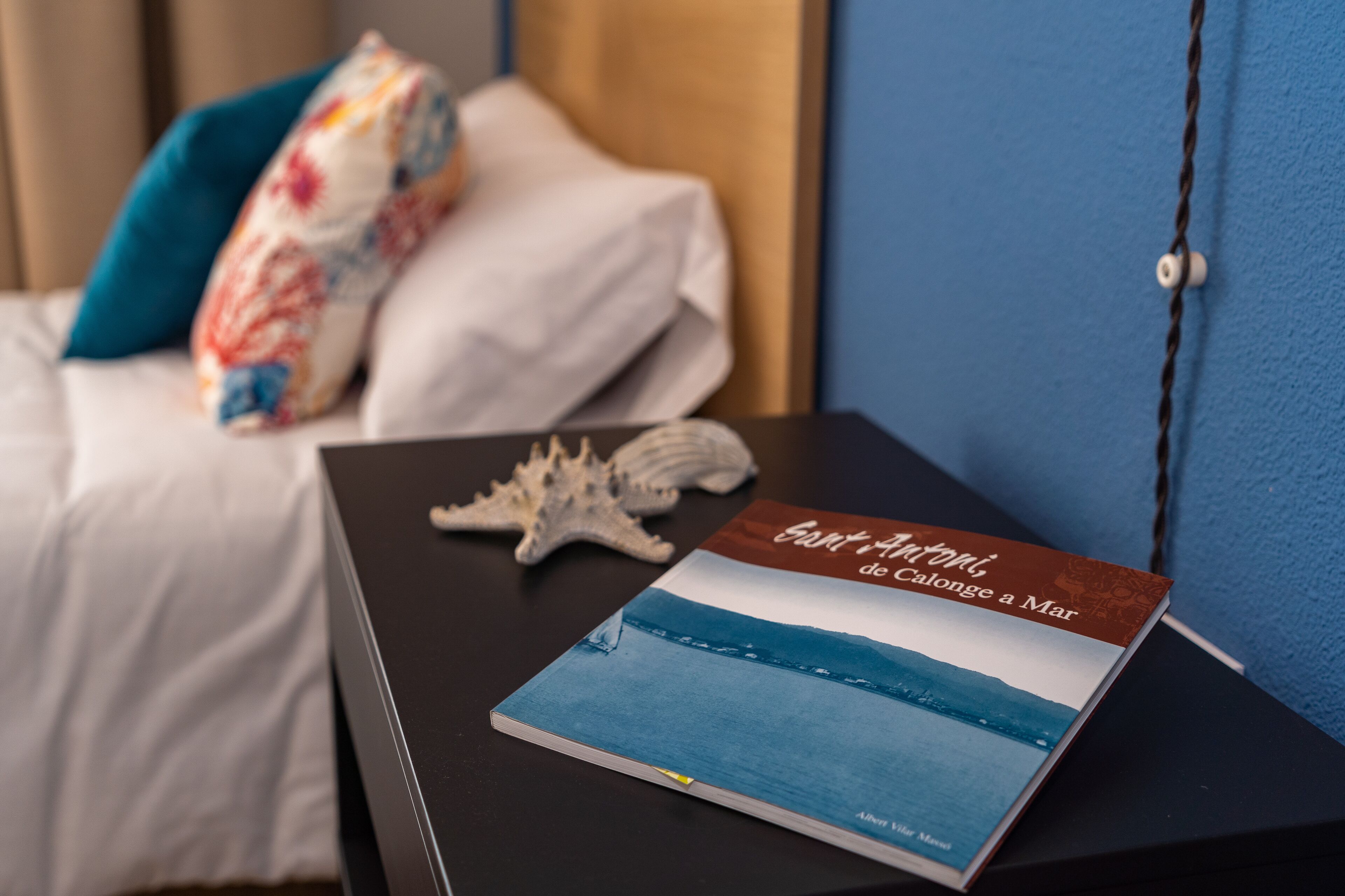 Een hotelkamer met een bed, een nachtkastje met een boek getiteld 'Santorini de Calonge a Mar' en een zeester.