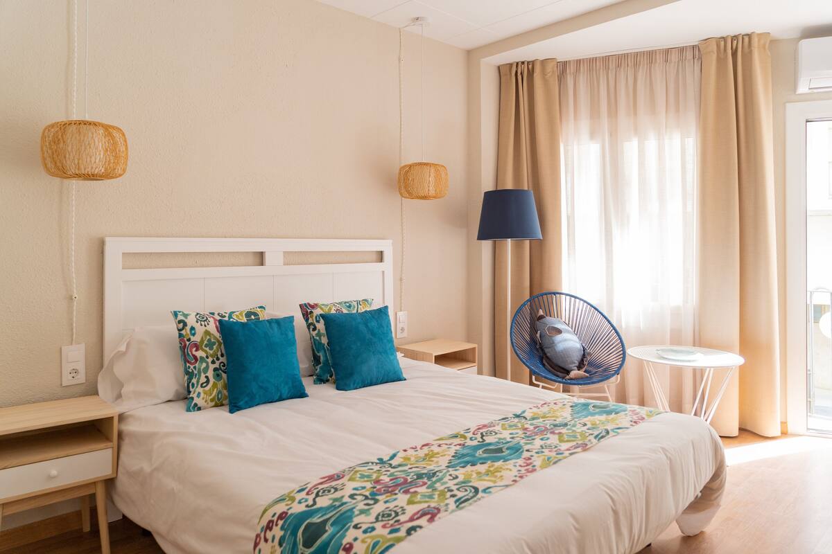 Une chambre d’hôtel bien aménagée, avec un grand lit, deux tables de chevet, une chaise, une petite table et une fenêtre avec des rideaux.