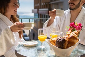 A couple enjoying breakfast on a balcony with a view of the city, a glass table, and a bowl of croissants and orange juice.