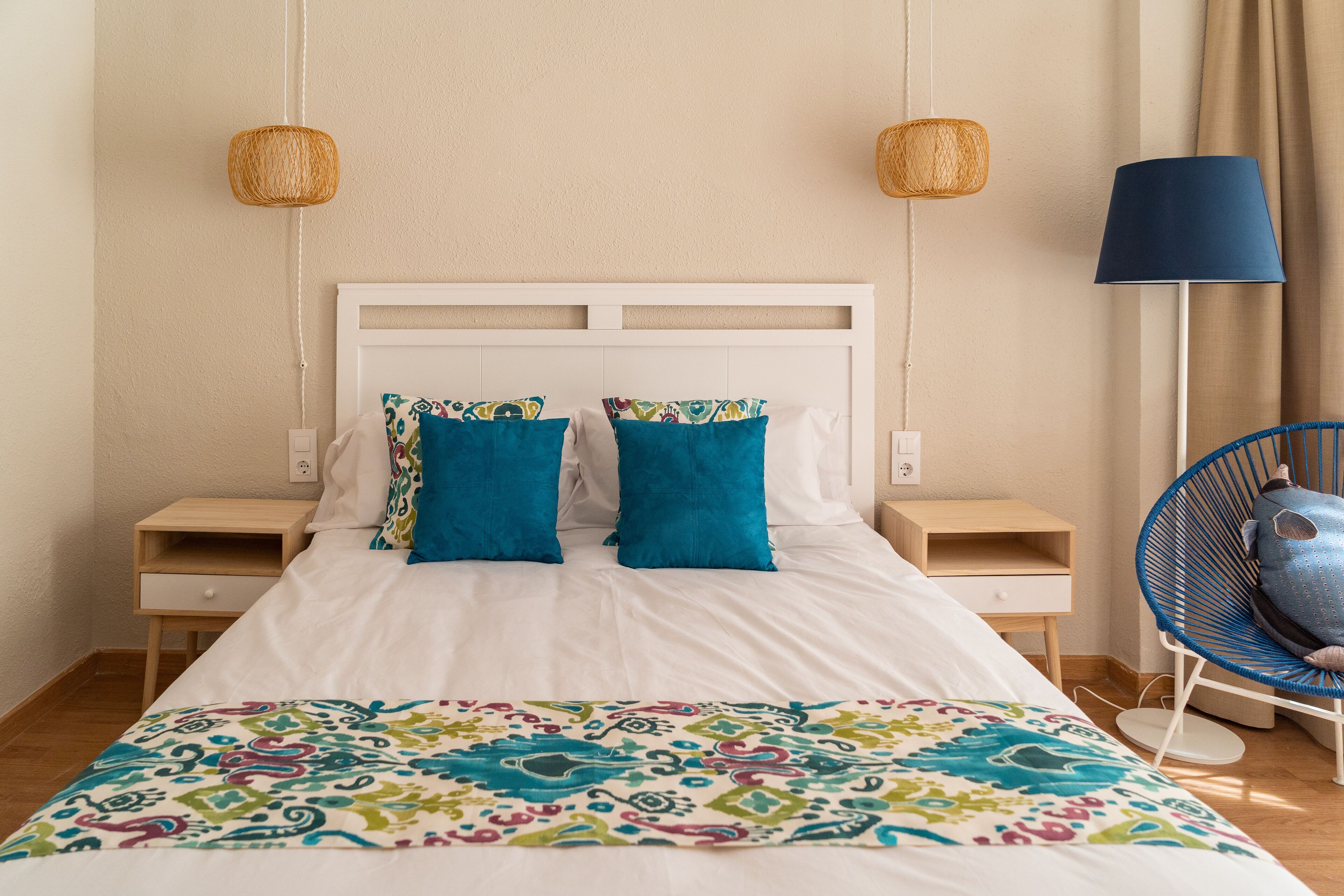 Un letto ben rifatto con una coperta colorata, due cuscini coordinati, due comodini, una poltrona blu e due lampade da parete in vimini.
