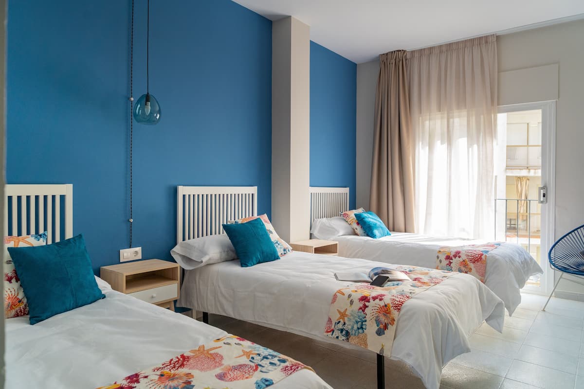 Une chambre d’hôtel avec trois lits, chacun doté d’une tête de lit blanche et d’oreillers bleus. Une table de chevet en bois se trouve entre les lits, et une chaise bleue est placée dans un coin de la pièce.