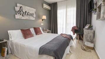 Un dormitorio con una cama grande, una mesita de noche con lámpara, un televisor montado en la pared y una ventana con cortinas.
