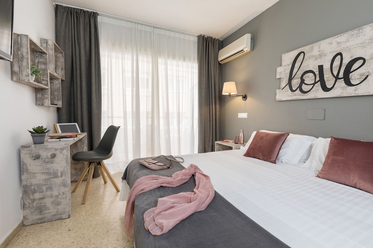 A hotel room with a bed, a desk with a chair, a potted plant, and a wall-mounted air conditioner.