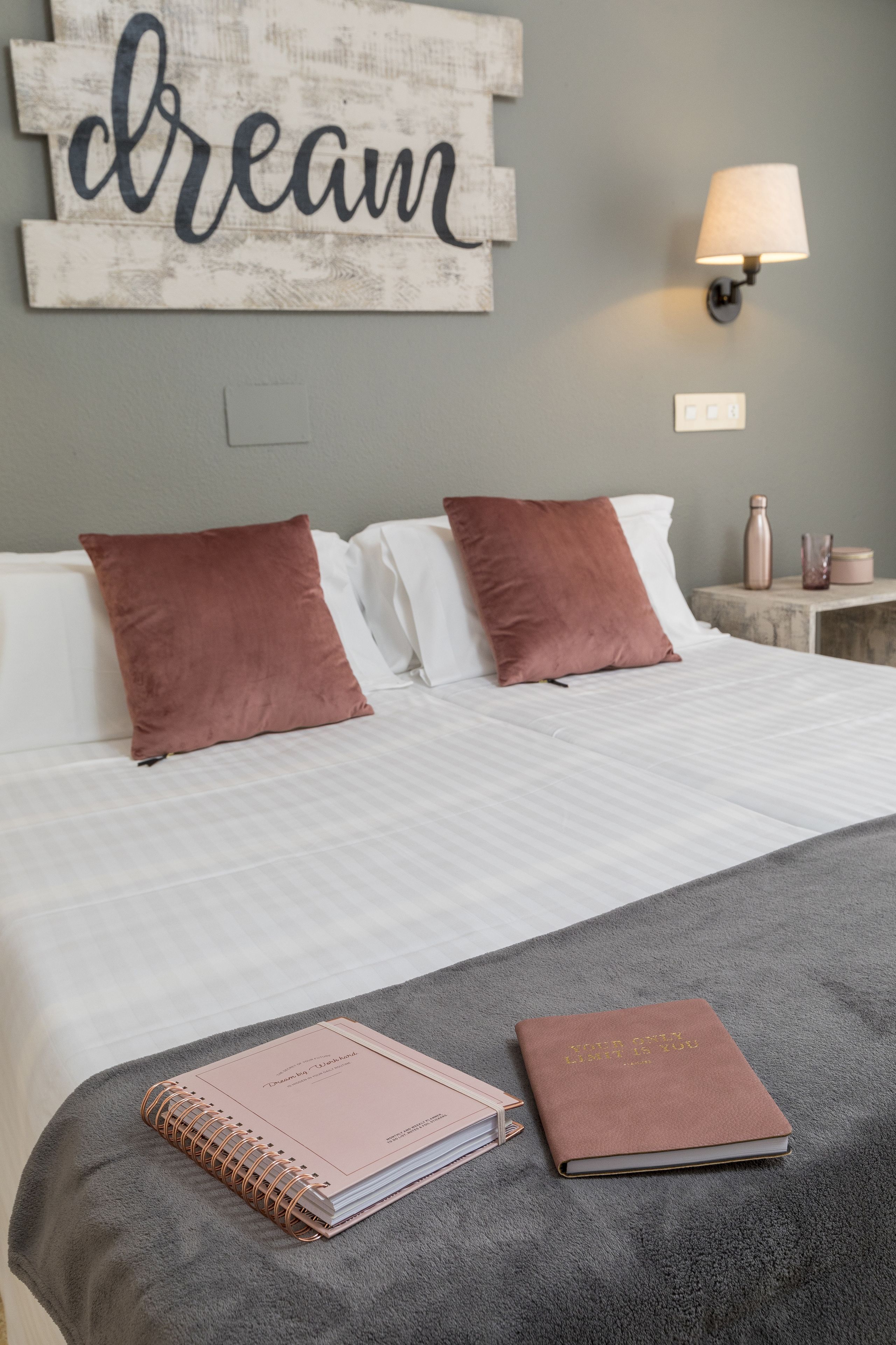 Una cama bien hecha con dos almohadas, una lámpara de noche, una mesita con una vela y un cuaderno sobre la cama.