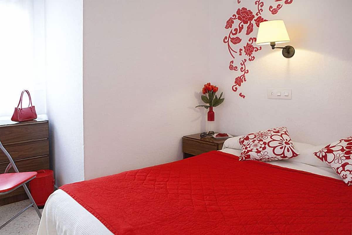 A bedroom with a red bedspread, a nightstand with a lamp, a dresser, a chair, and a window with a red handbag on it.