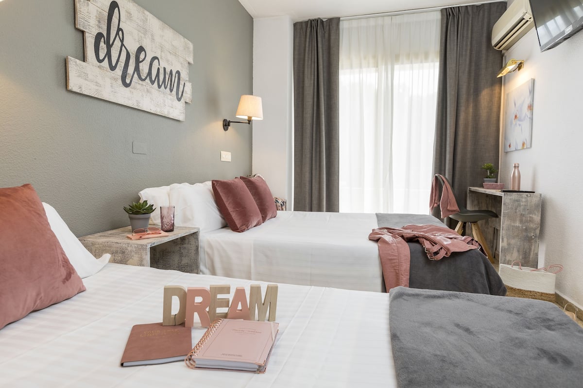 A hotel room with two beds, a wall-mounted lamp, a large window with curtains, and a 'dream' sign on the wall.