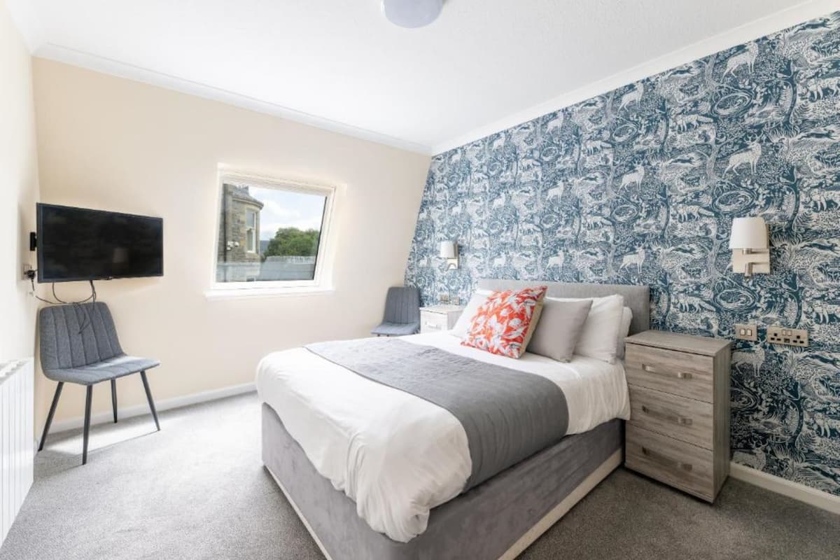 A bedroom with a bed, a chair, a TV, a window, and patterned wallpaper.