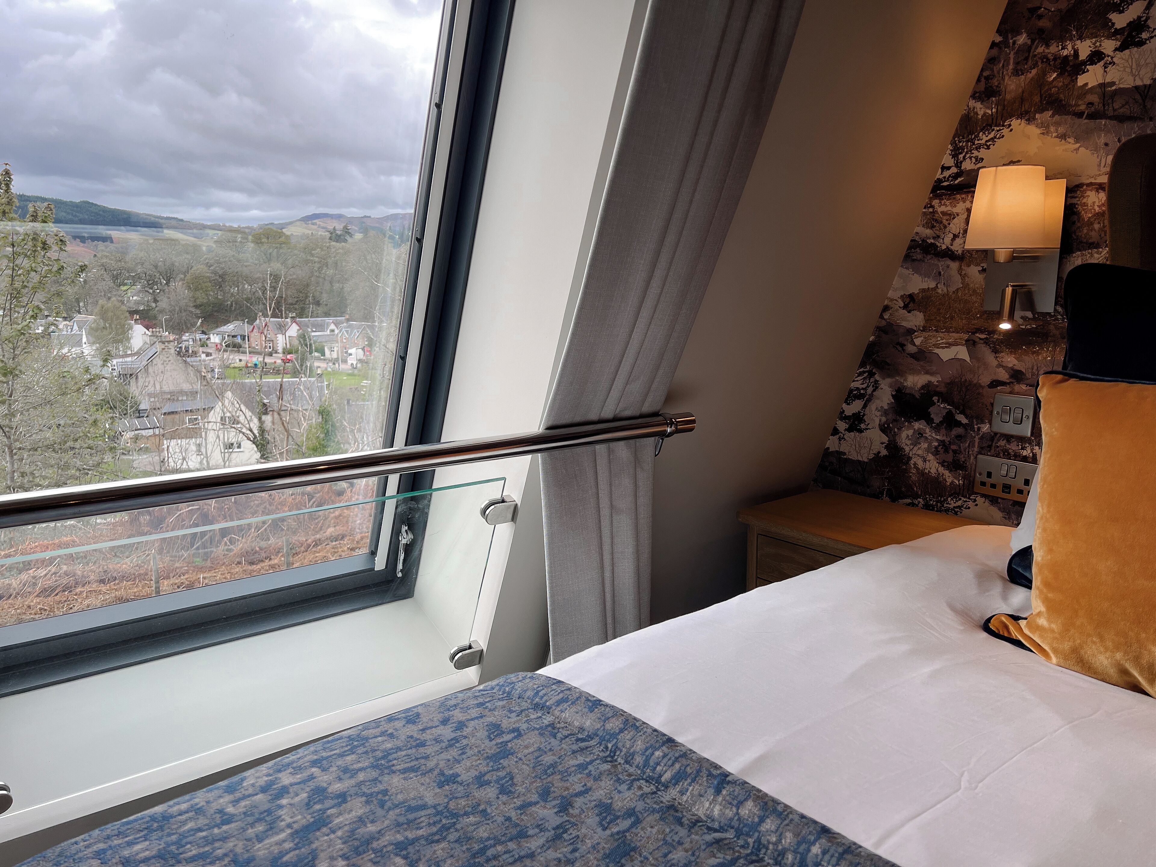 A hotel room with a bed, a window with a view of a town, a lamp, and a wall with a textured pattern.