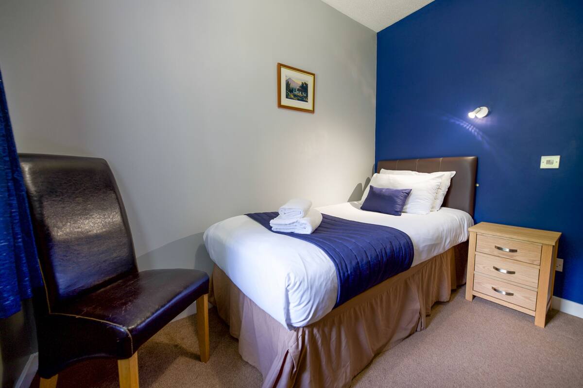 A neatly arranged bedroom with a bed, a chair, a bedside table, and a wall-mounted light.