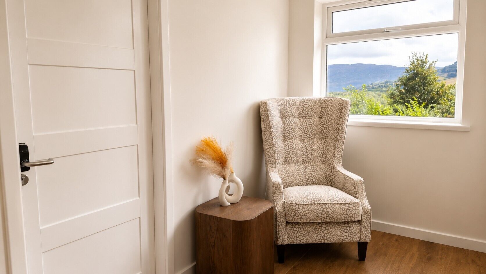 A room with a white door, a wooden side table with a vase, and a patterned armchair.