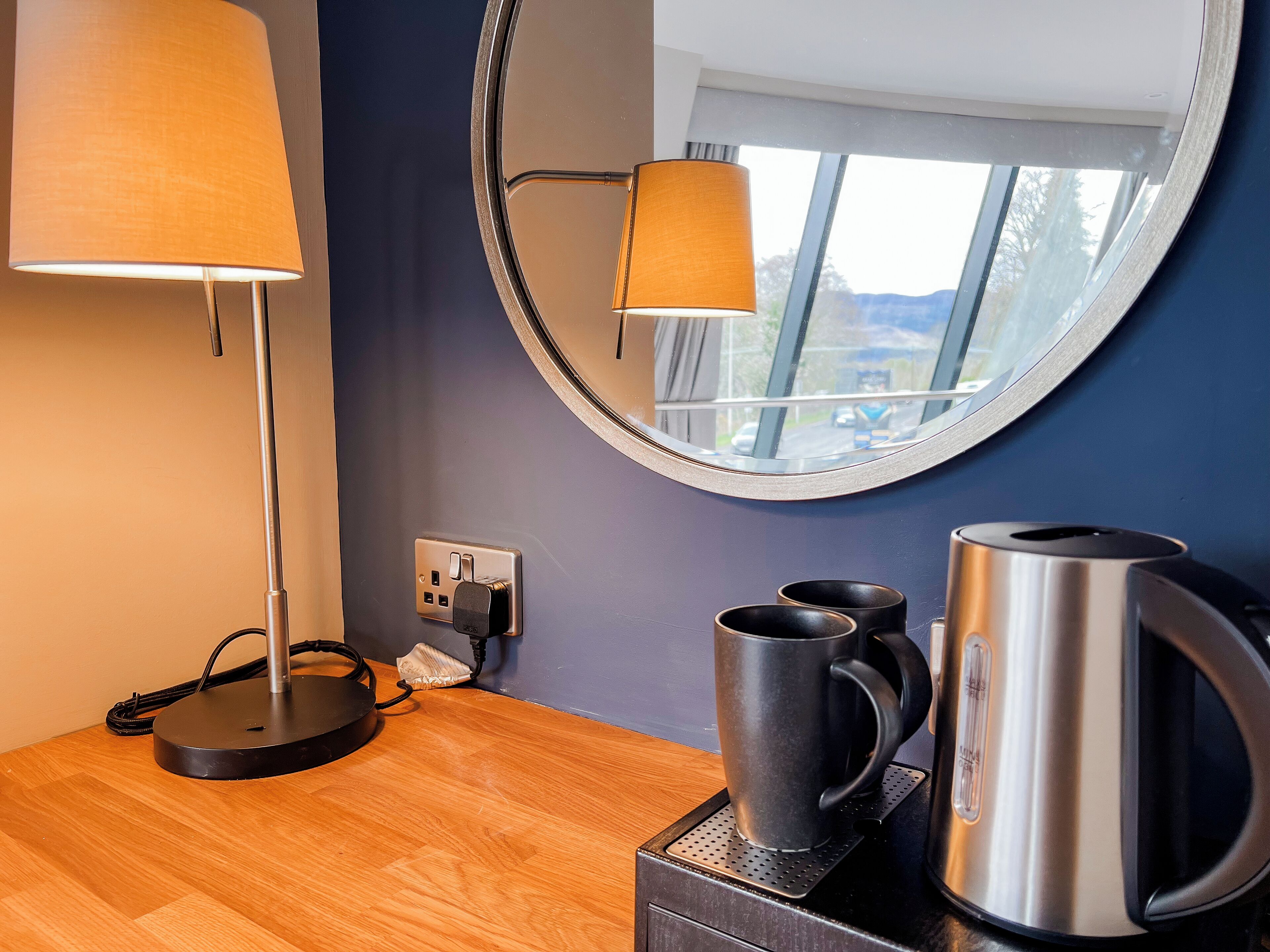 Une chambre d’hôtel avec un plancher en bois, un miroir rond, une lampe, deux tasses et une bouilloire posée sur une petite table.