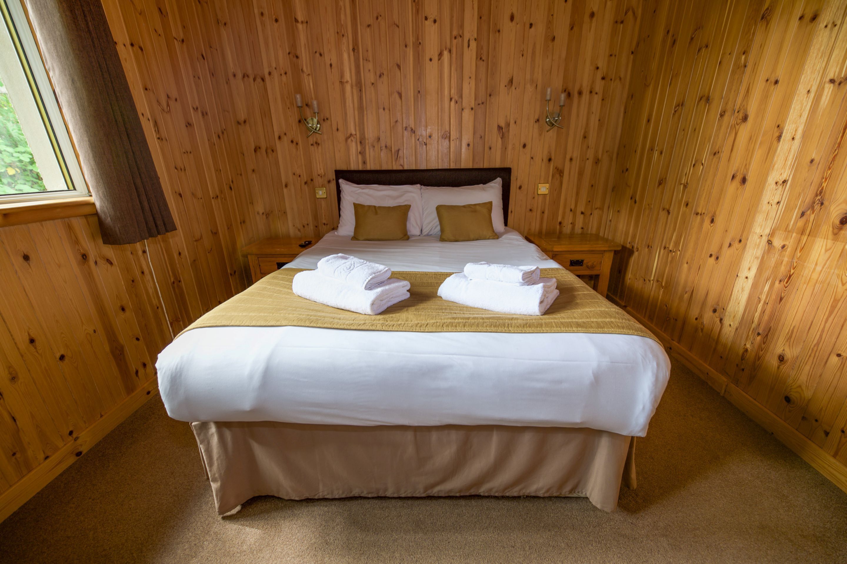 A neatly made bed with white linens and two gold pillows, a wooden headboard, and a window with curtains.