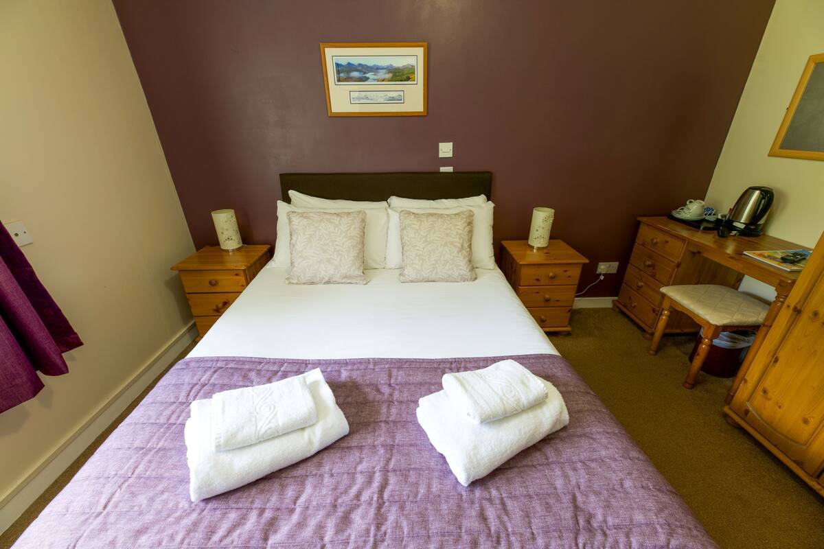 A neatly made bed with a purple bedspread and two towels, a wooden nightstand with a lamp, a wooden dresser, and a desk with a chair.