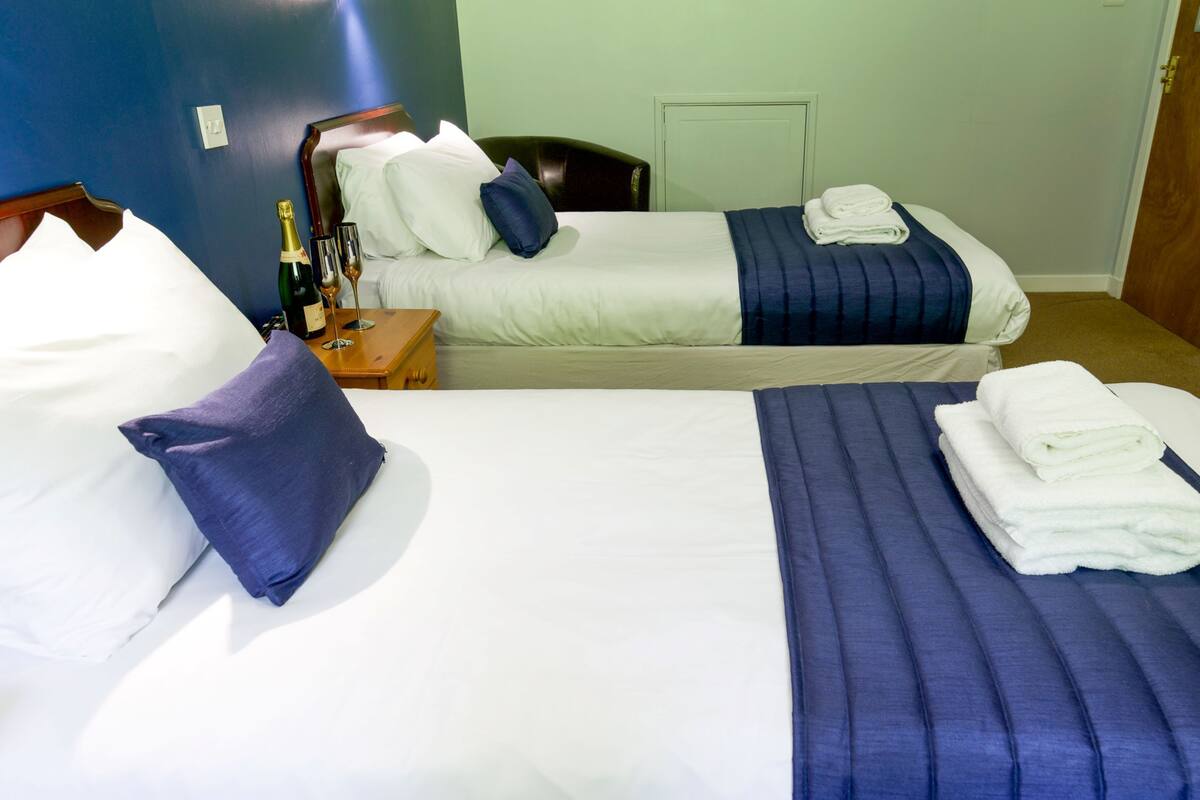 A hotel room with two beds, each with a different color bedspread, a nightstand with a bottle and glasses, and a door to the right.